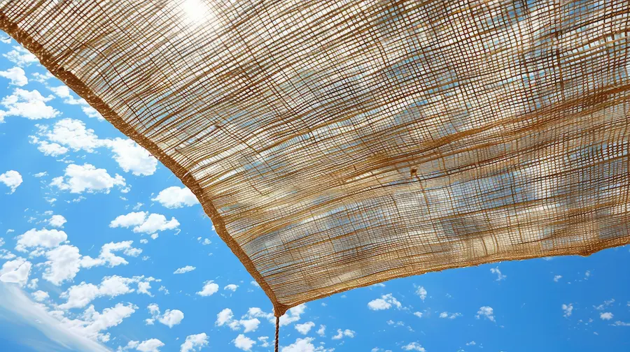 青空の下で強い日差しを遮る、ナチュラルな質感のオーニング（シェード）。夏のウッドデッキやテラスの暑さ対策・日よけとして設置された、おしゃれで機能的なエクステリアのイメージ。