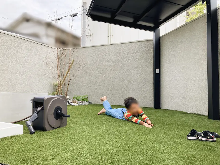 日よけ対策として設置された独立型テラス屋根。外壁に固定せず自立するため、建物への影響を抑えながら人工芝の庭に広い日陰を作る施工事例。シェードよりも耐久性が高く、子供の遊び場としても最適。