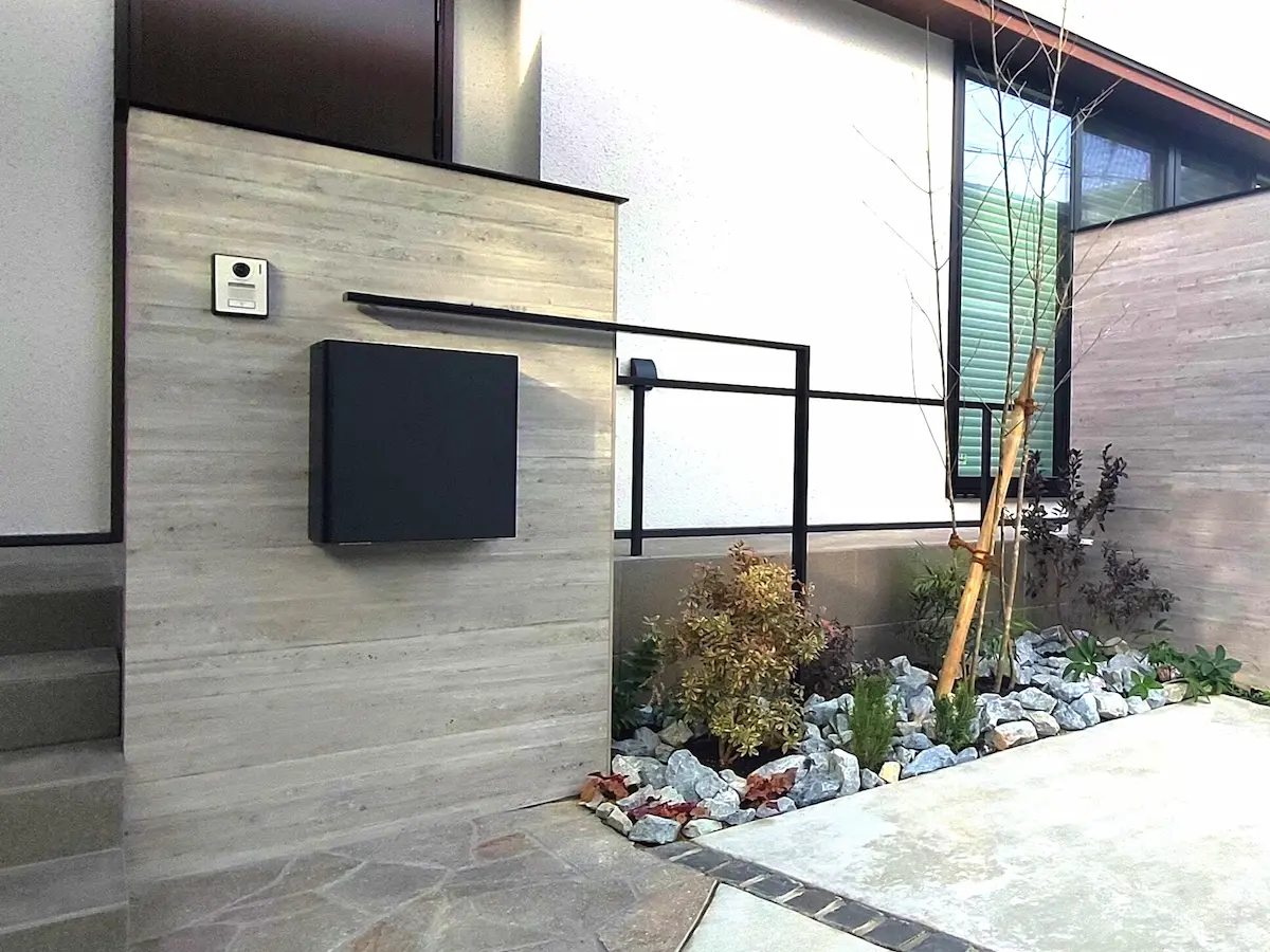 モダンな住宅の外構デザイン。杉板目調のコンクリート壁、黒いポストとインターホン、植栽スペースにはグリ石（割栗石）と低木が配置されている