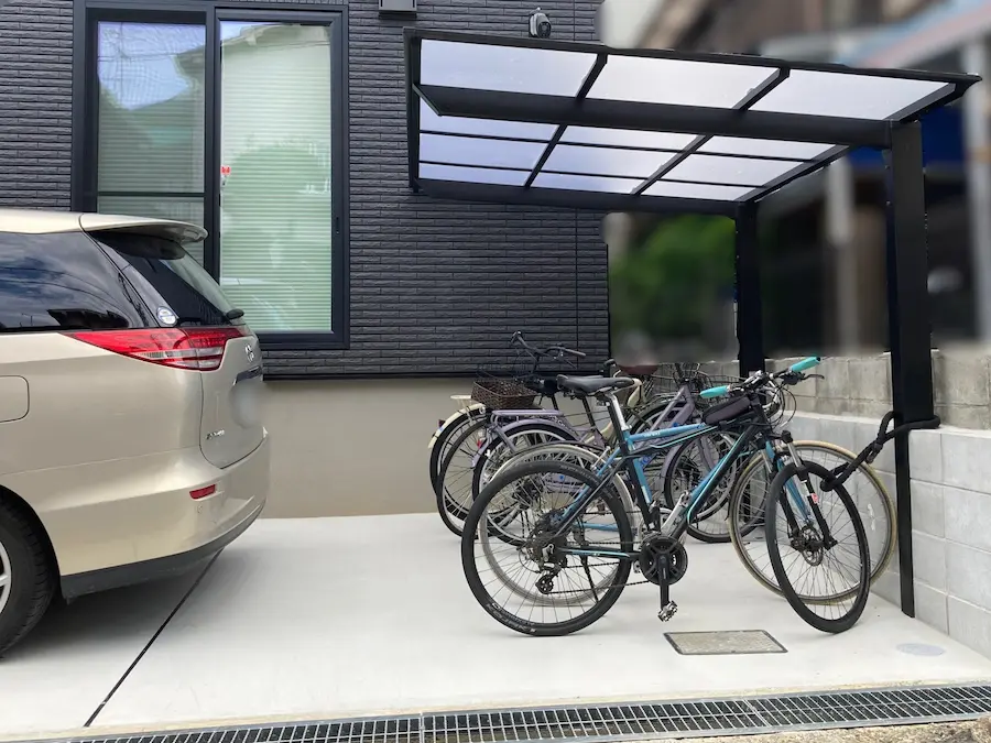 新築戸建ての土間コンクリート駐車場に設置された、おしゃれな黒フレームの片流れカーポート（サイクルポート）。マウンテンバイクやシティサイクルなど複数台の自転車を雨から守る、機能的で省スペースな駐輪スペースの施工事例。