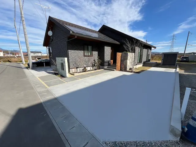 SOTORIE群馬伊勢崎のエクステリア施工実績5、黒いモダンな住宅の全景。広い土間コンクリートの駐車場と、玄関前の目隠しとして配置された石積み風のモルタル造形ウォールがおしゃれな外構。