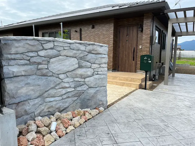 SOTORIE群馬伊勢崎のエクステリア施工実績6、落ち着いたグレー系の石積み風モルタル造形ウォール。門塀の中央には家主の名前が刻まれ、足元にはブラウン系の石が置かれた外構デザイン。独立型の緑のポストがレンガ風の建物と統一感を保っている