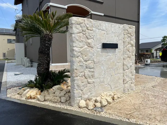 SOTORIE群馬伊勢崎のエクステリア施工実績7、明るいベージュ系のゴツゴツとした石積み風モルタル造形門柱。黒い表札とソテツ、白い石を組み合わせたリゾート風外構。